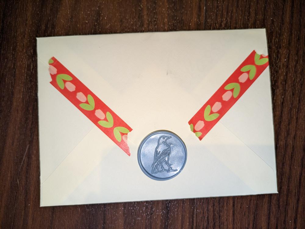 A white 5x7 envelope made with thin woven linen paper. There is a sage blue wax seal with a crow design on it. There is also washi tape sealing the envelope with tulips on it